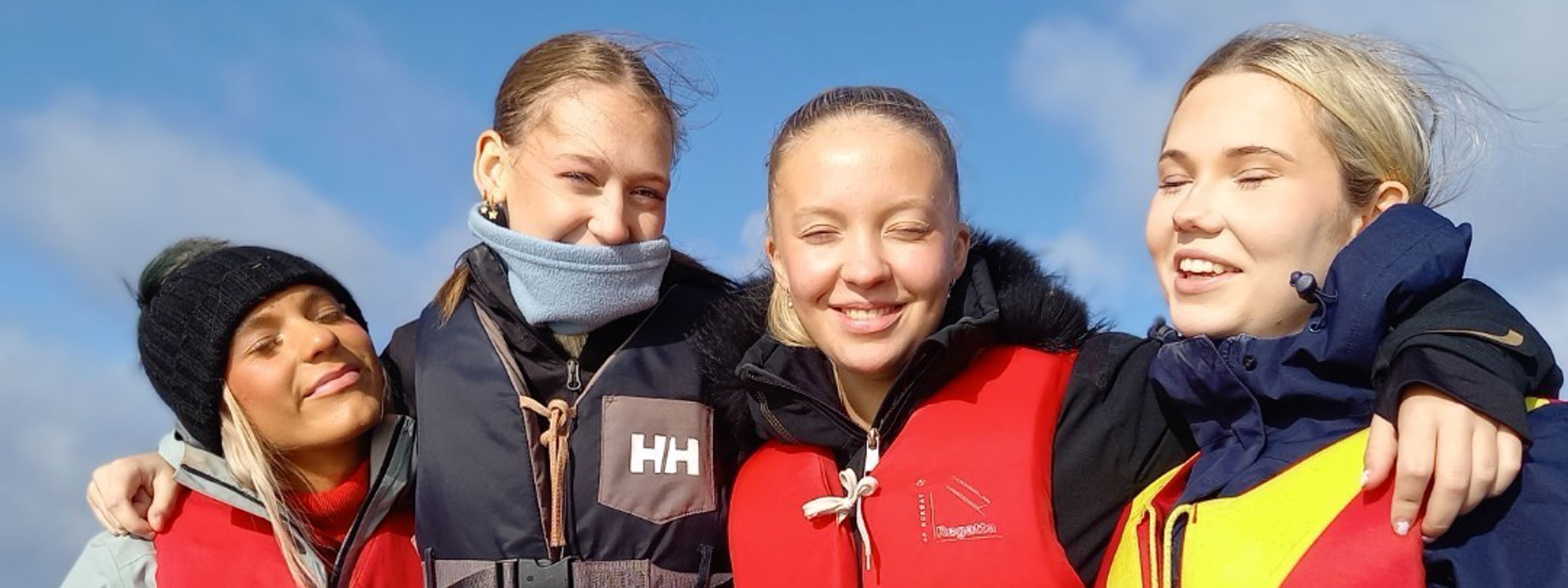 4 jenter som står tett saman med redningsvestar på