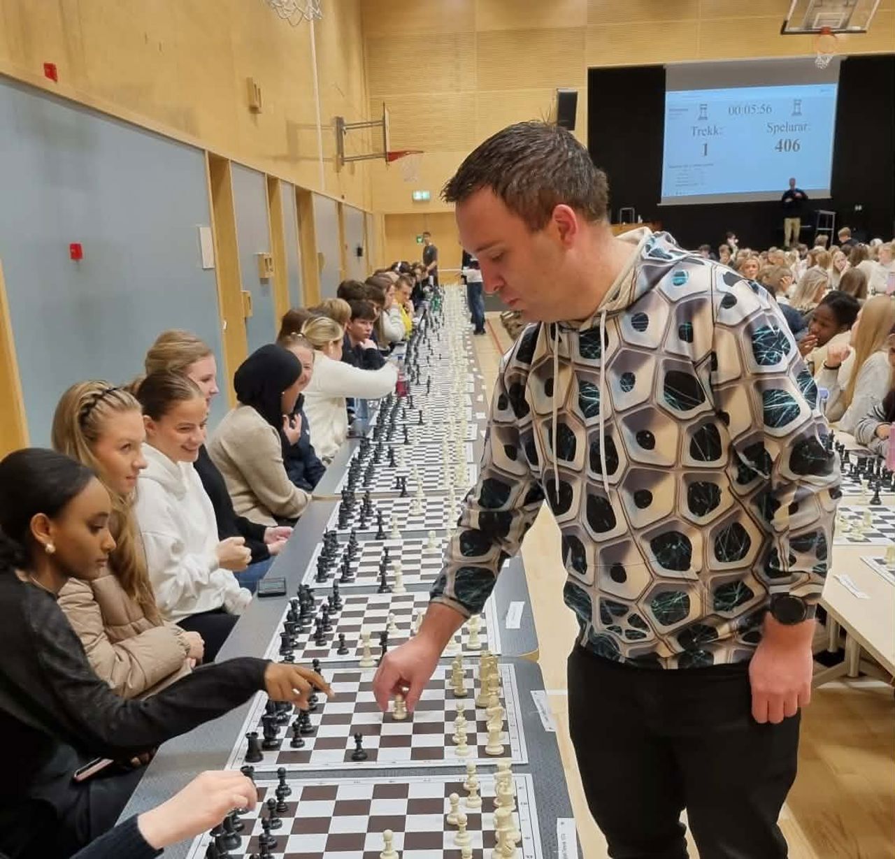 En sjakk spelar går langs eit lanbord med mange sjakk brett på og spelar med elevane
