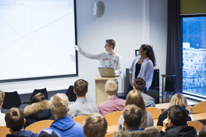 To elevar står saman framfor ei undervisningstavle, elevar som sit og føljer med.