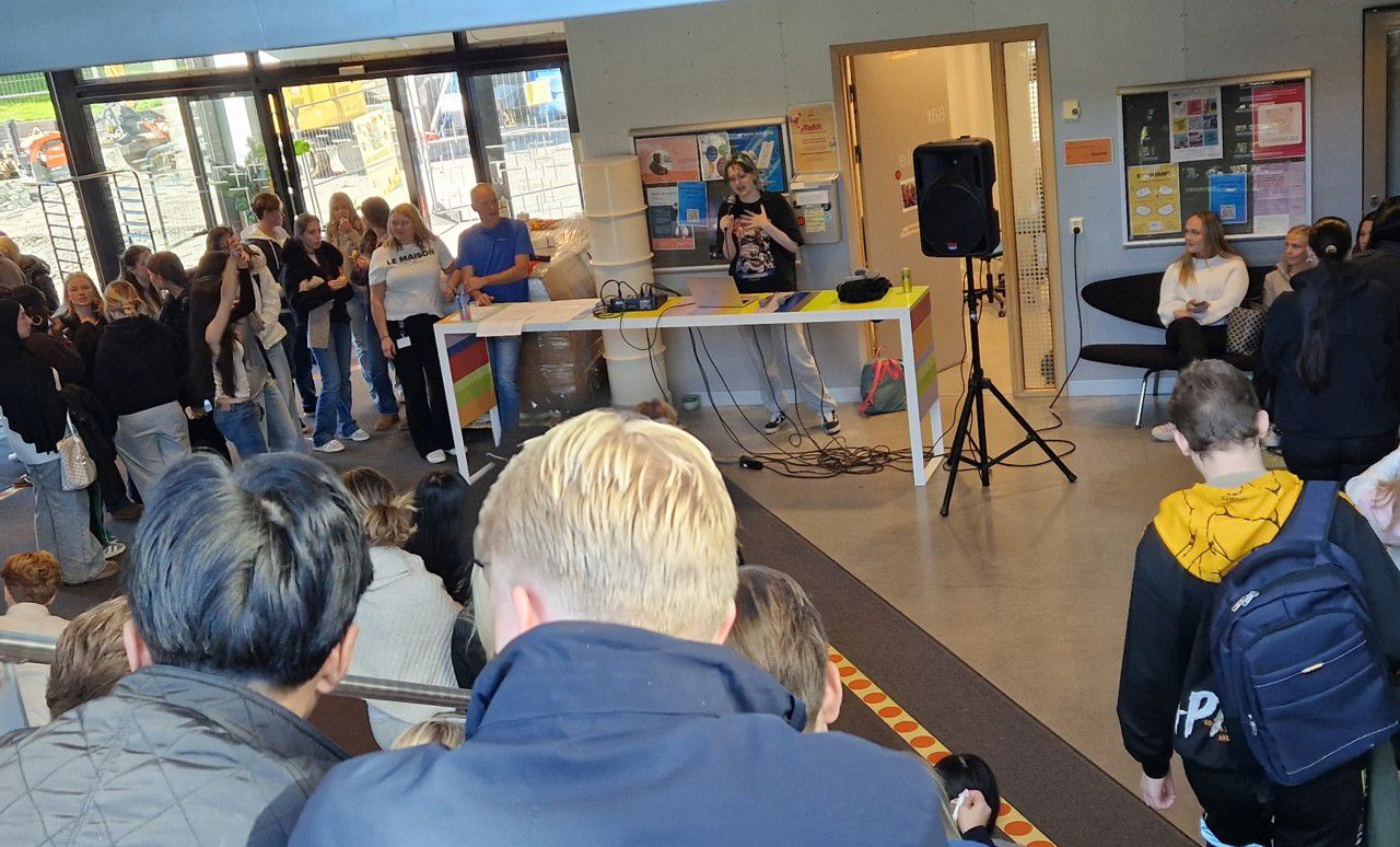 Mange elevar sit og står rundt ei jente som syng karaoke