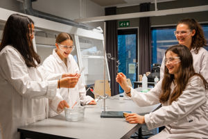 4 jenter i kvite frakkar sit rundt eit bord og gjera eit kjemisk forsøk