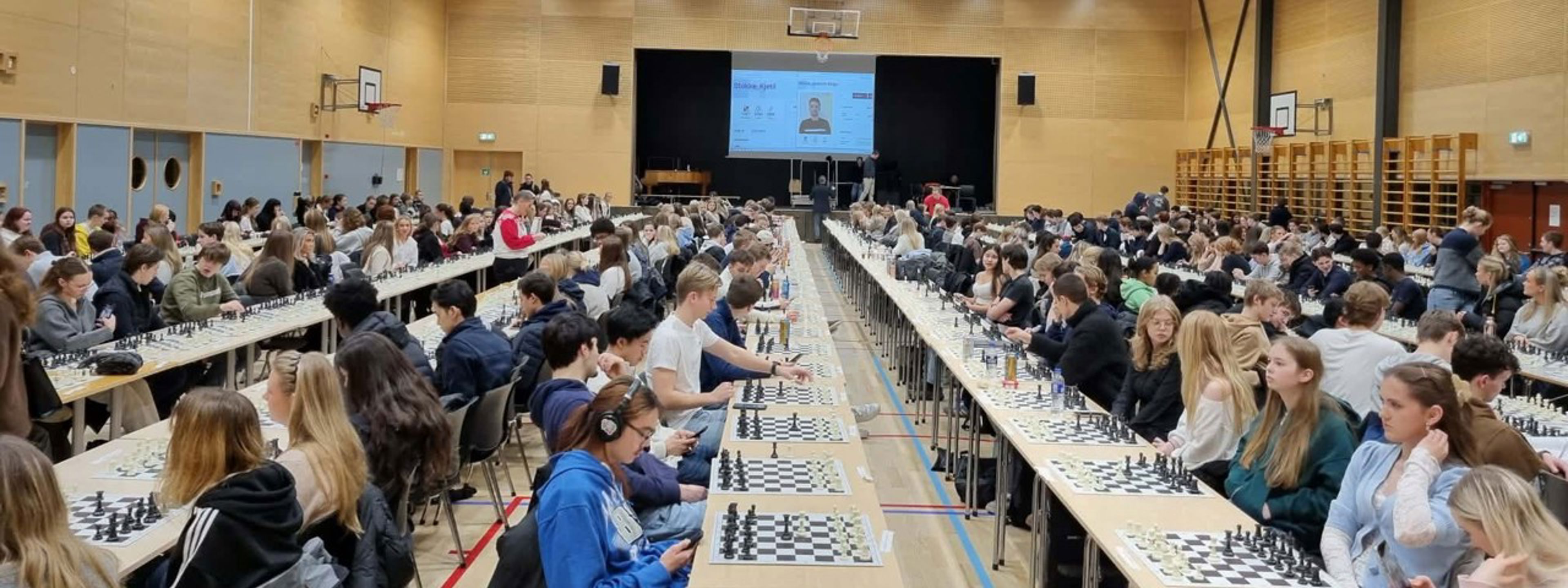 Ein gymsal full av elevar som sit ved langbord og speler simultansjakk