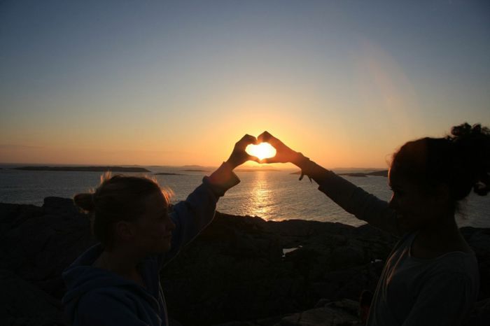 To jenter held hendene saman og lagar eit hjarte i solnedgang i havet